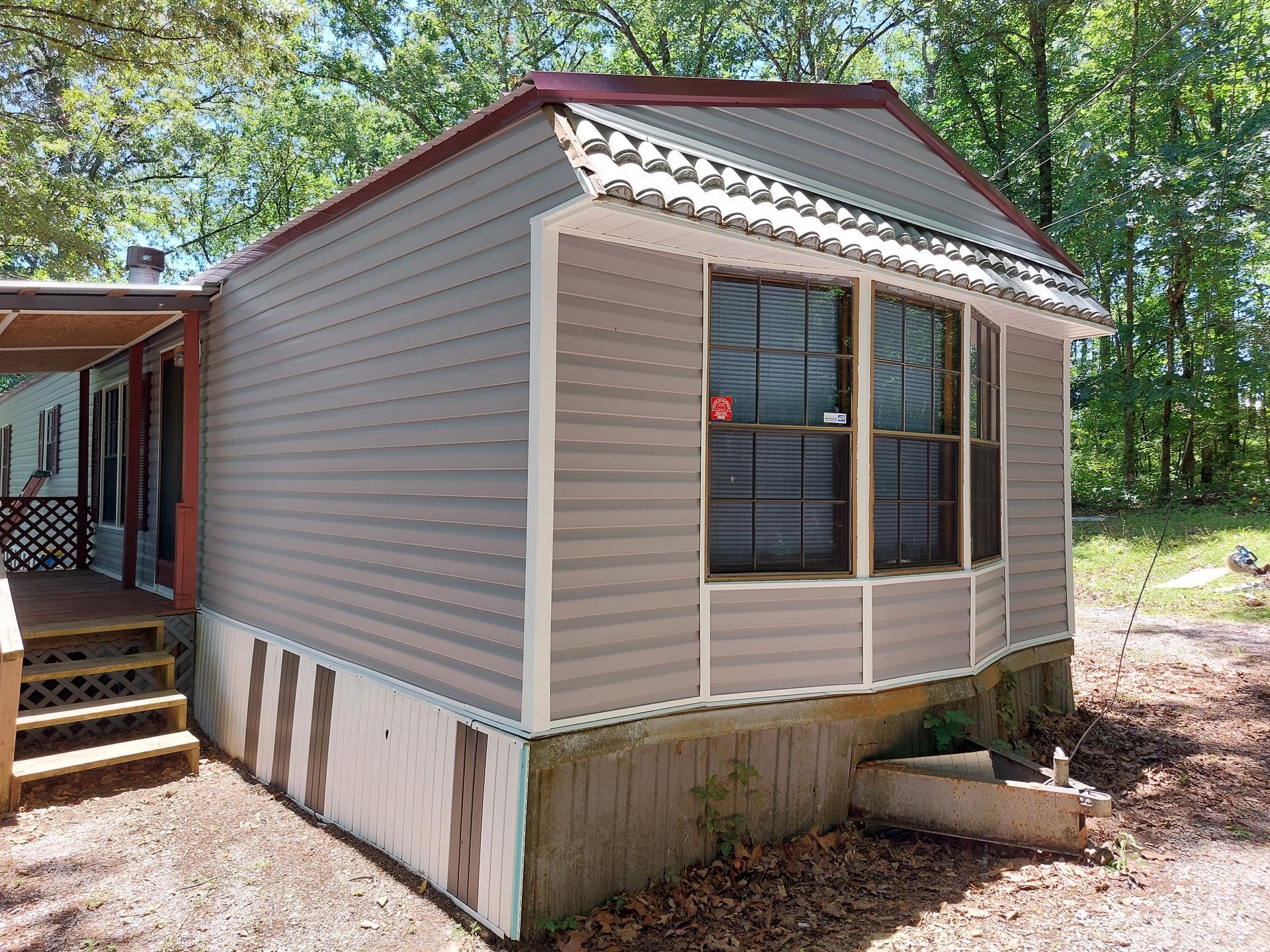 Small beige mobile home with red roof trim and windows, elevated on blocks in a wooded yard.