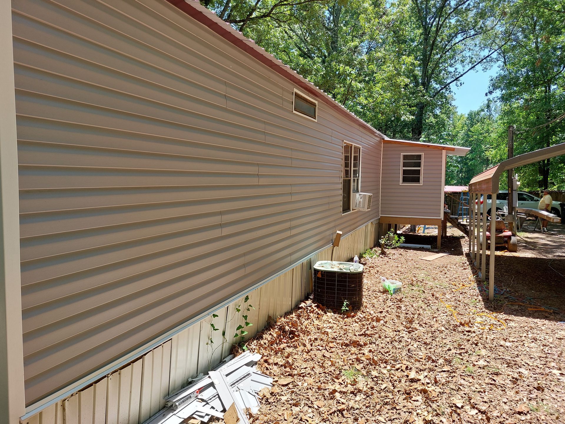 Side of tan mobile home with white skirting, dirt yard, and a small shed in the background.
