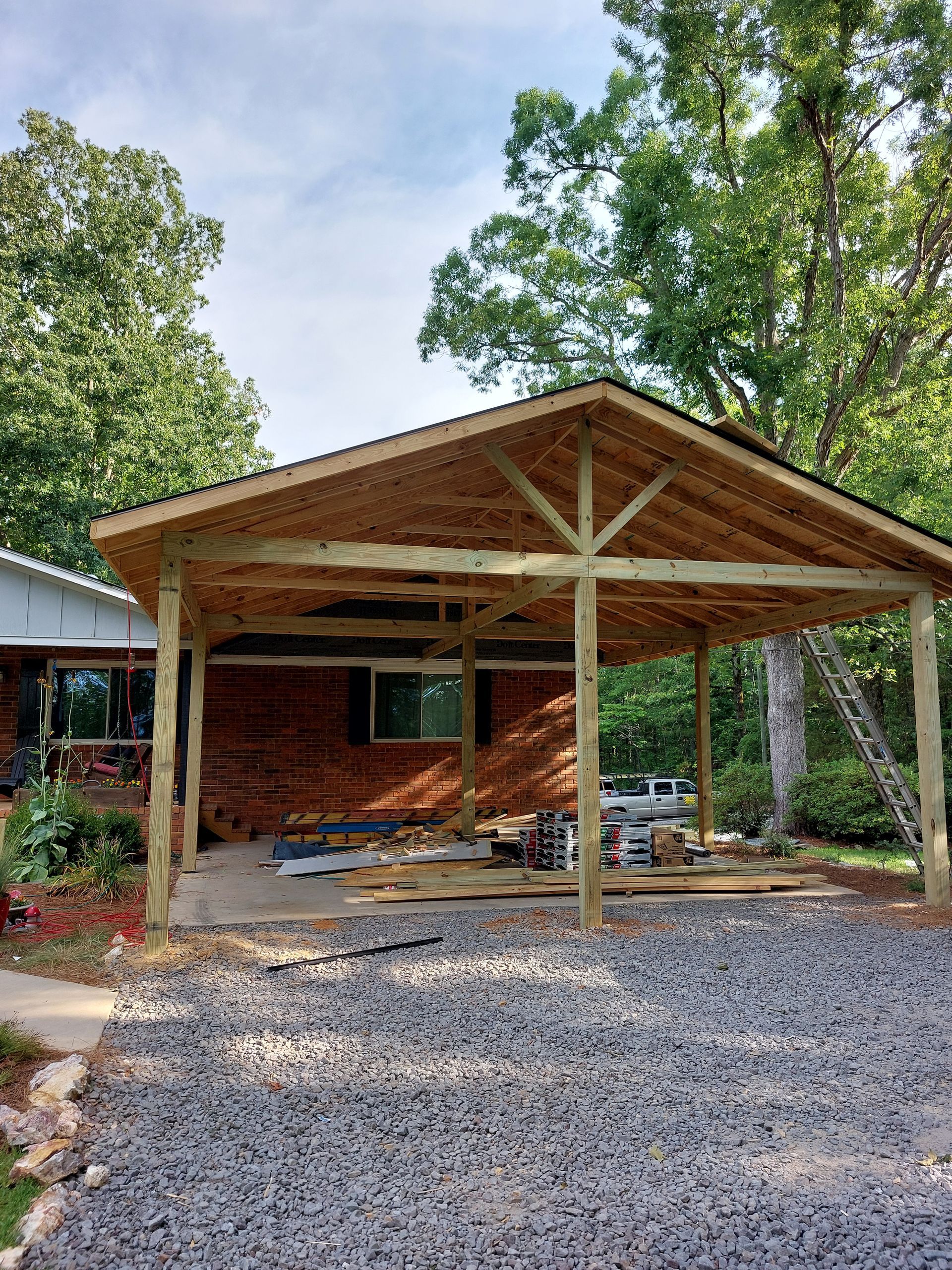 Wooden carport attached to a brick house, with gravel driveway and trees in the yard.