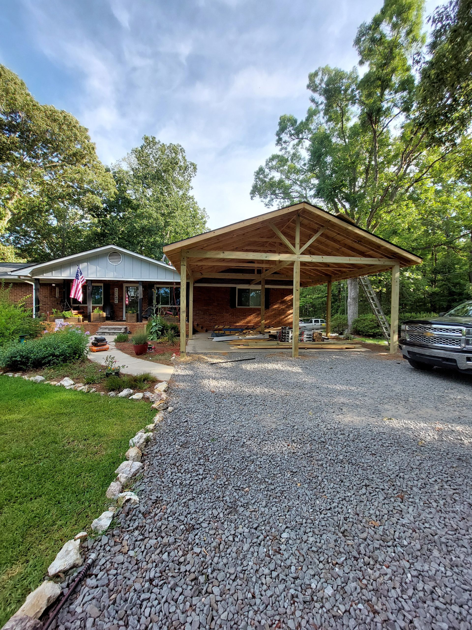 Gravel driveway leading to a wooden carport beside a house, with trees and a parked car.