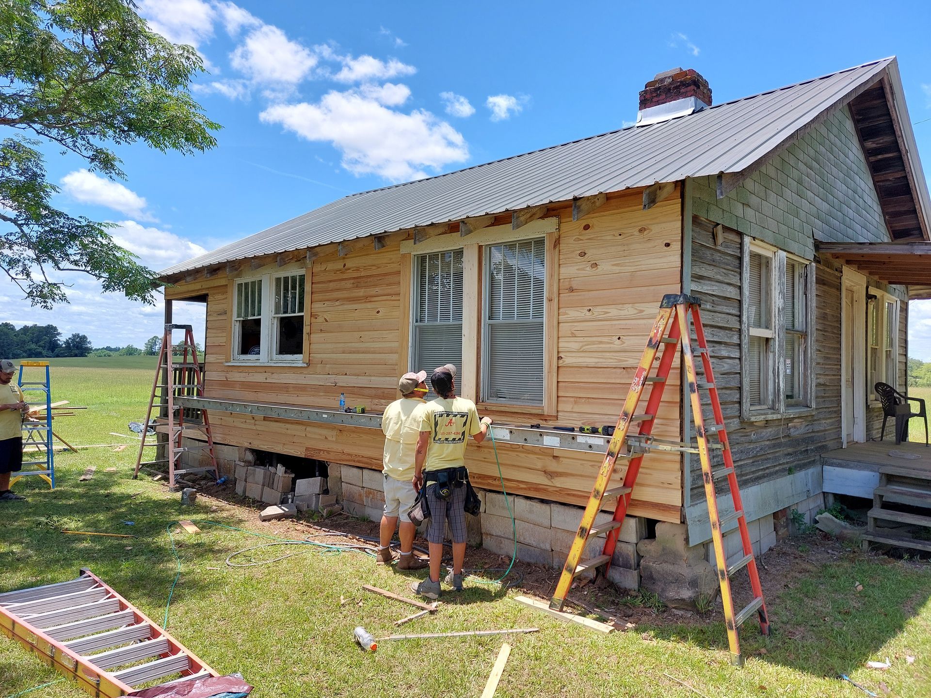 Workers installing siding on a small house with ladders and tools in a grassy yard.