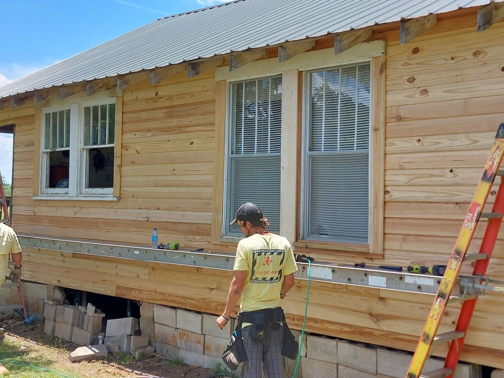 Worker siding a house under construction, with ladder and windows visible.