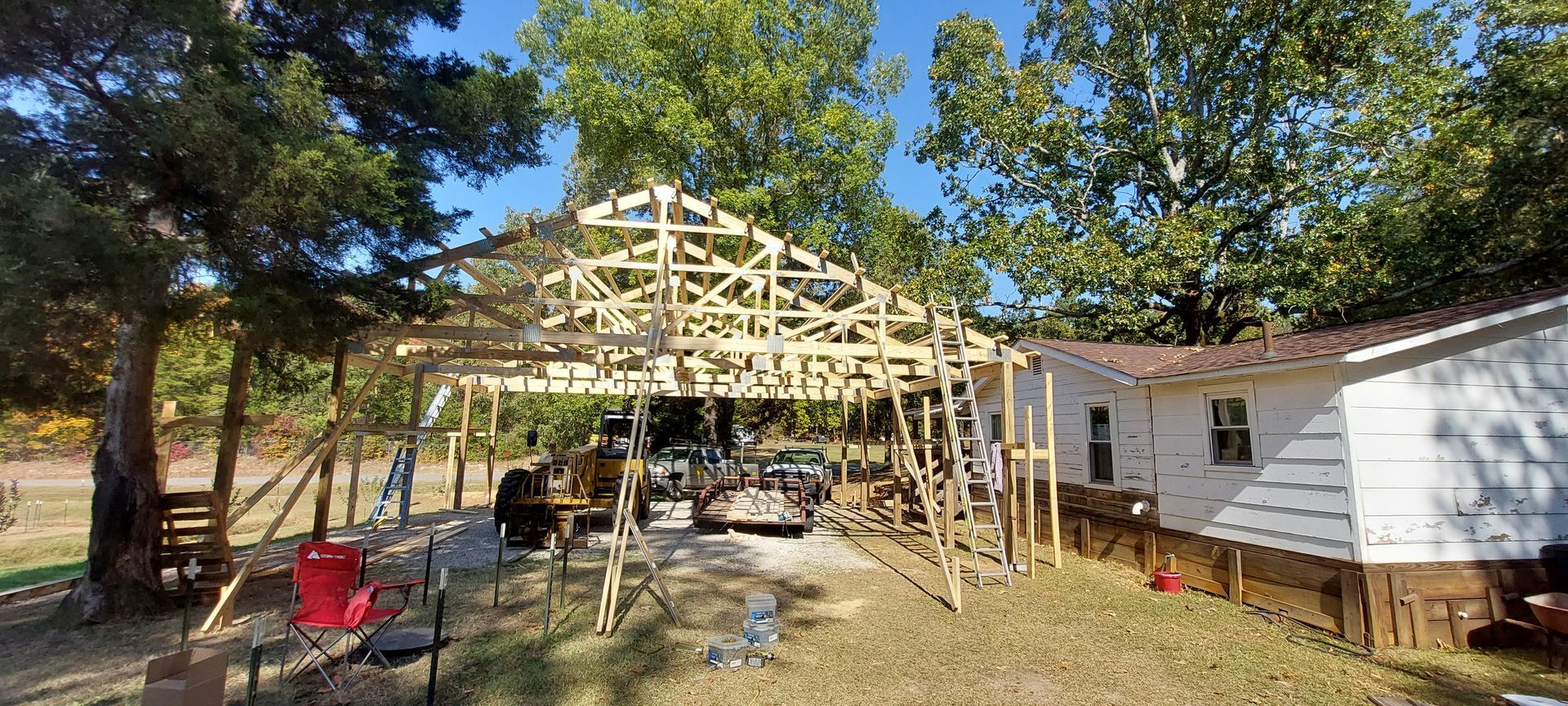 Wooden roof frame under construction beside a white mobile home in a wooded yard