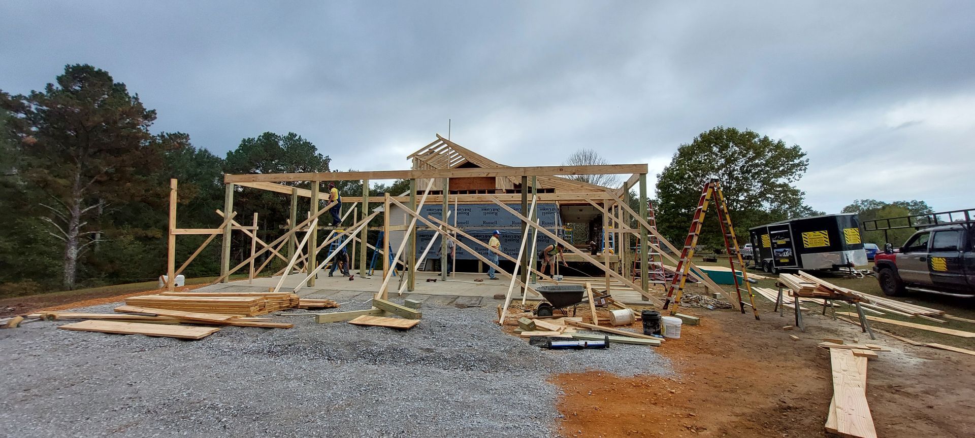 Wooden building frame under construction on a gravel lot, with lumber, equipment, and trees in the background