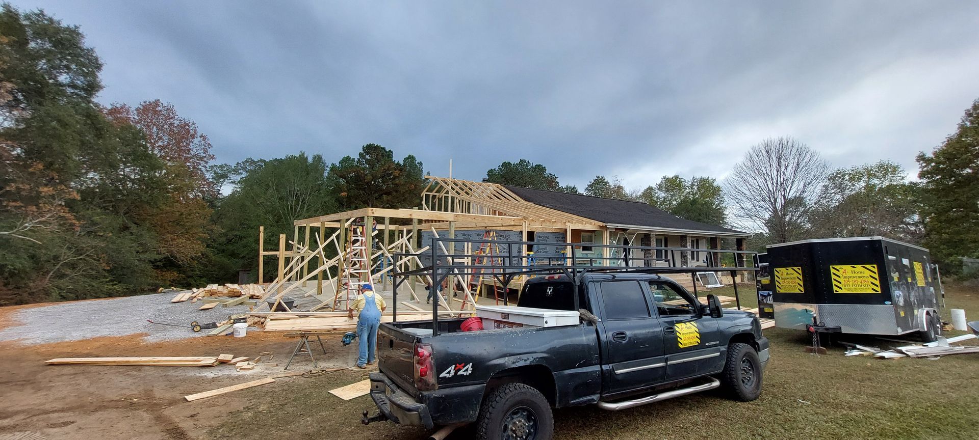 Construction site with wooden house frame, black work truck, and trailer on a dirt lot under cloudy skies