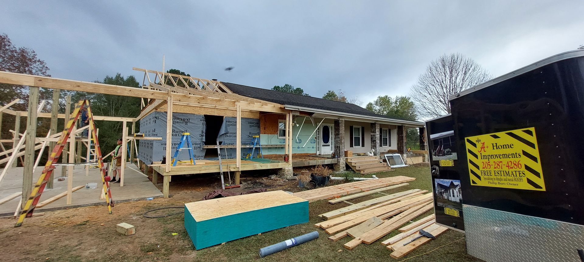 Partially built wooden house with exposed framing, lumber piles, and a dumpster in the foreground