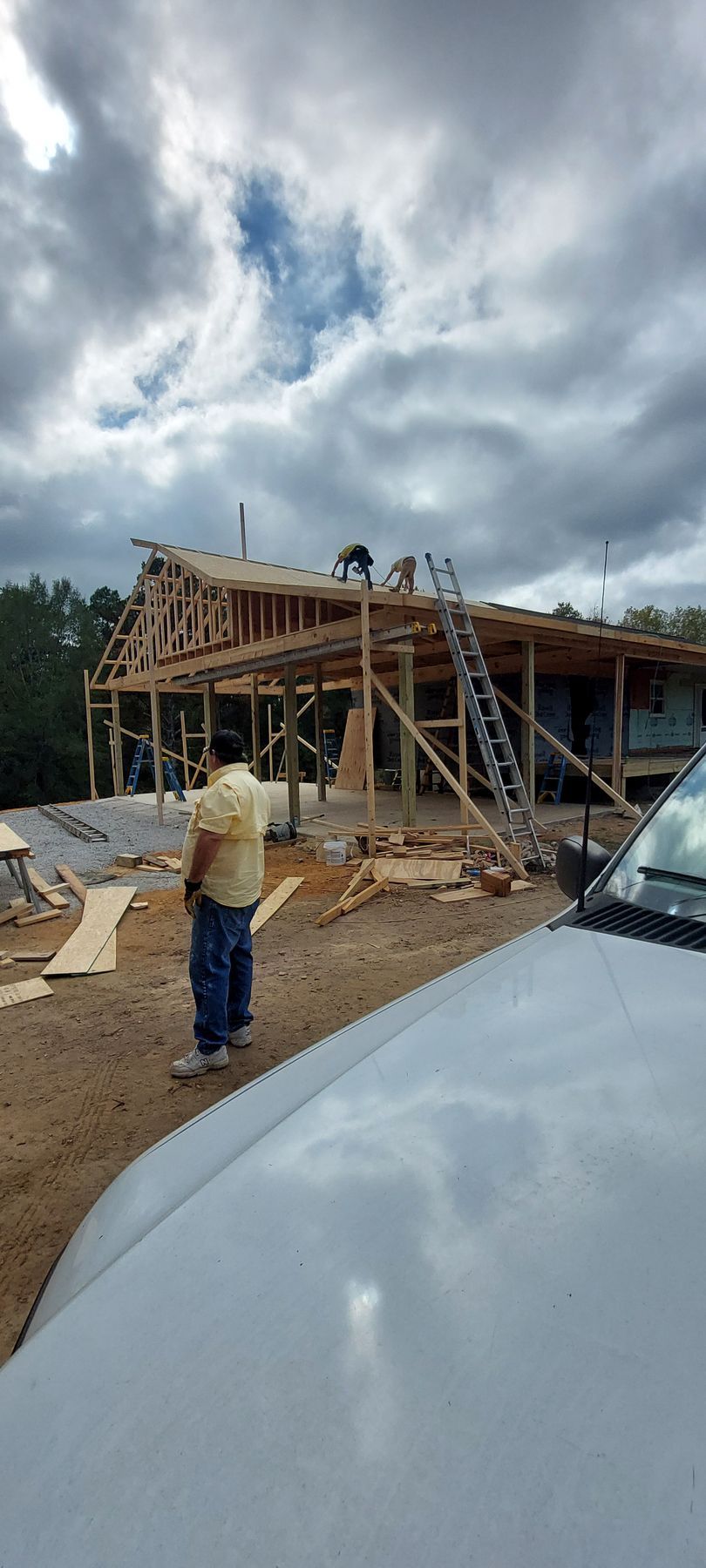 Partially built wooden house under cloudy sky, with a person standing nearby and a white vehicle in the foreground