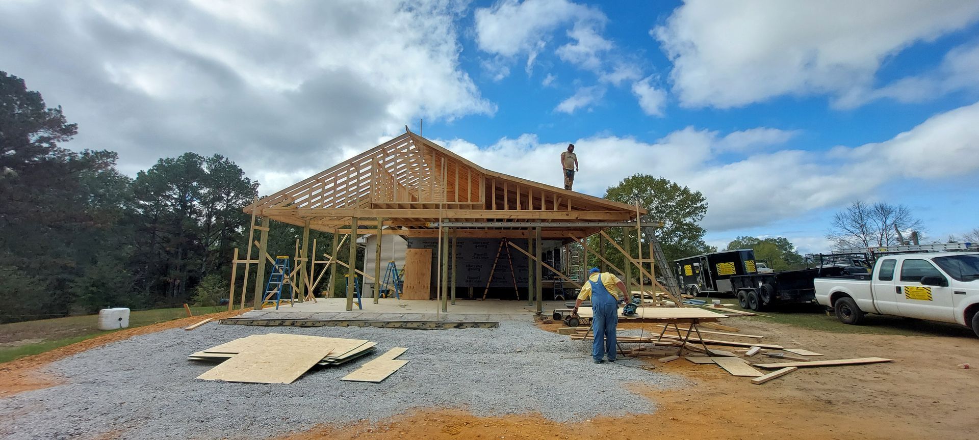 Wooden house under construction with roof framing, workers, and vehicles on a gravel lot under cloudy sky