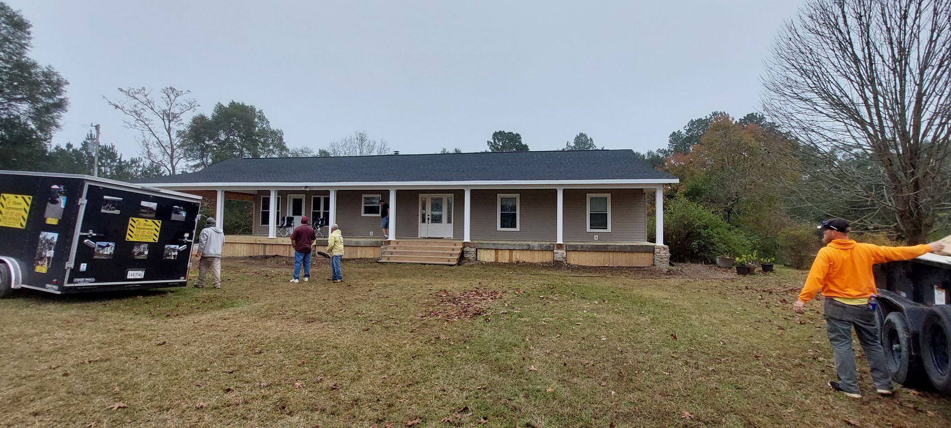 People standing in a yard outside a single-story brick house, with an overturned vehicle on the left.