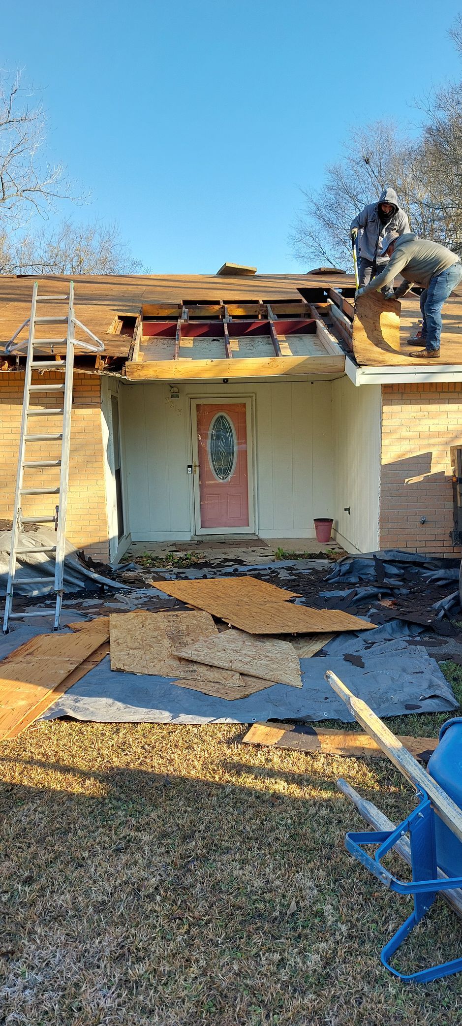 Person working on roof above a small house with a pink door, ladder at left, debris in front yard