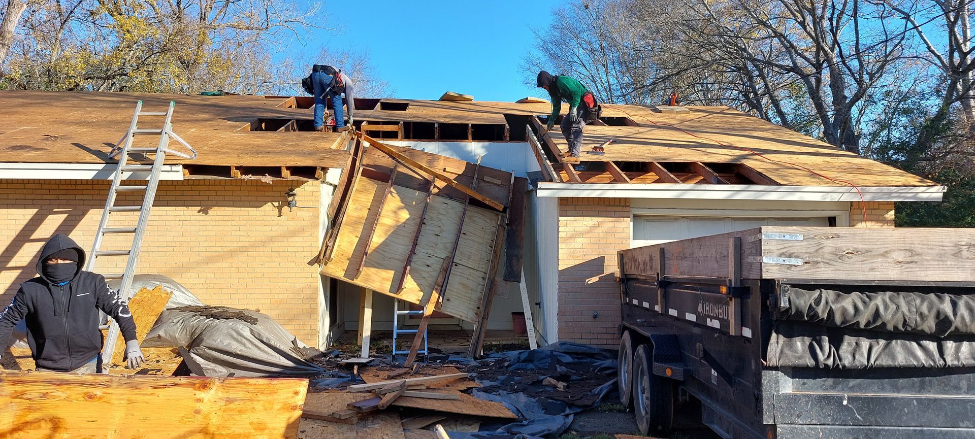Workers repairing a damaged roof on a house with ladders and wood debris under clear blue sky