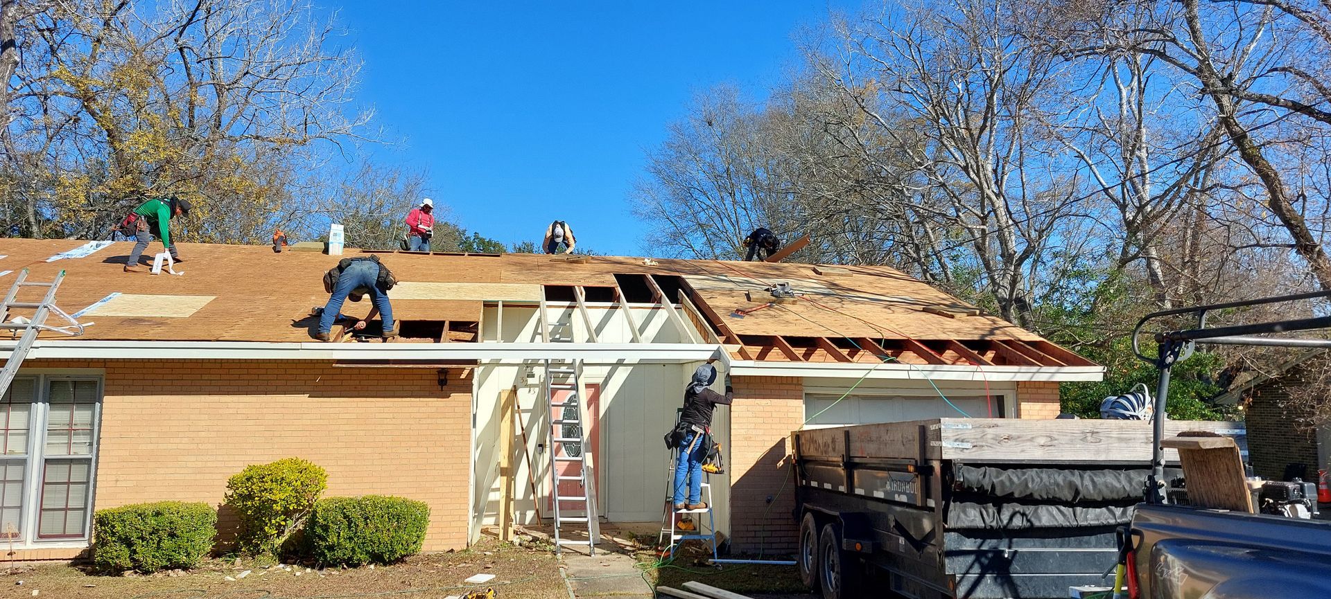 Workers installing a new roof on a house with a ladder and construction trailer in the yard