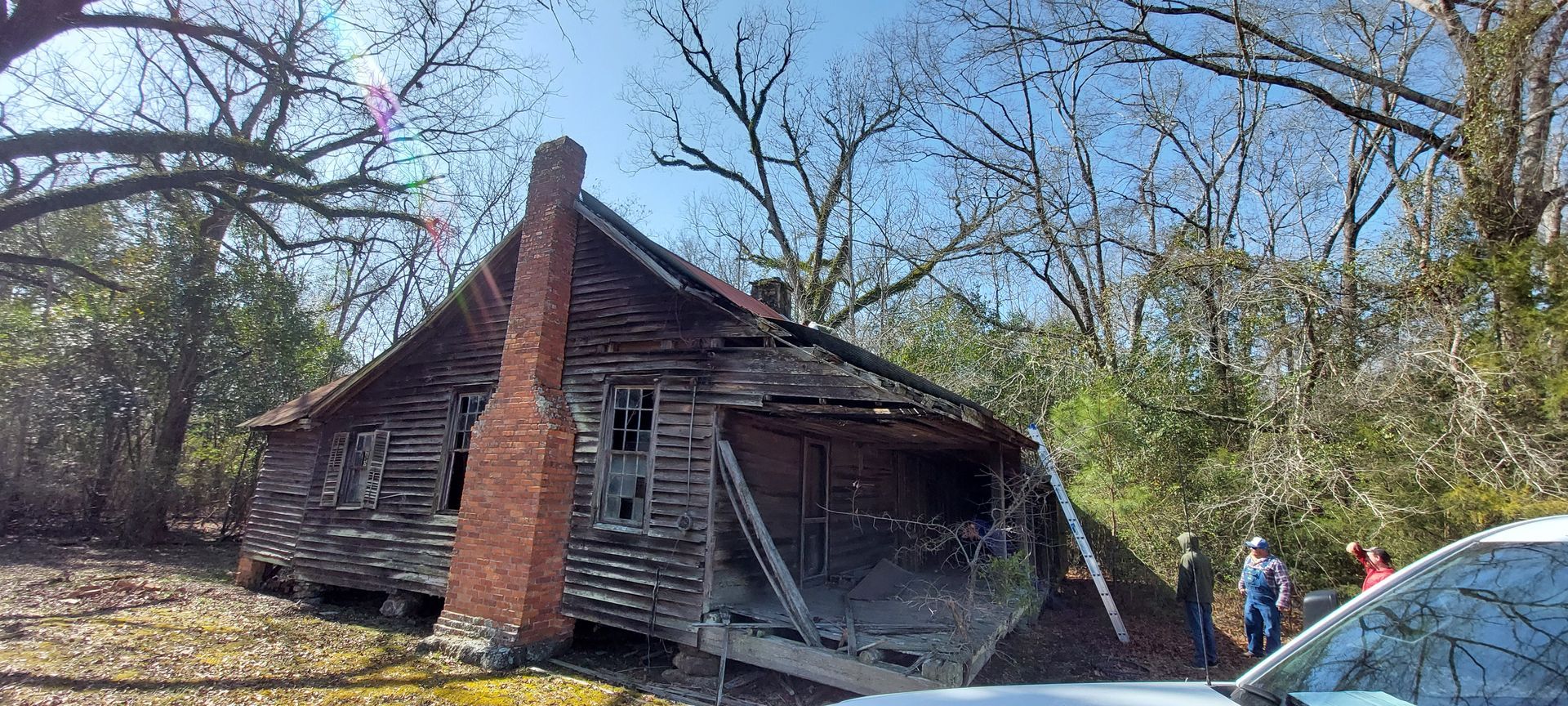 Crumbling old wooden house with brick chimney, trees in front, and a person near a parked car