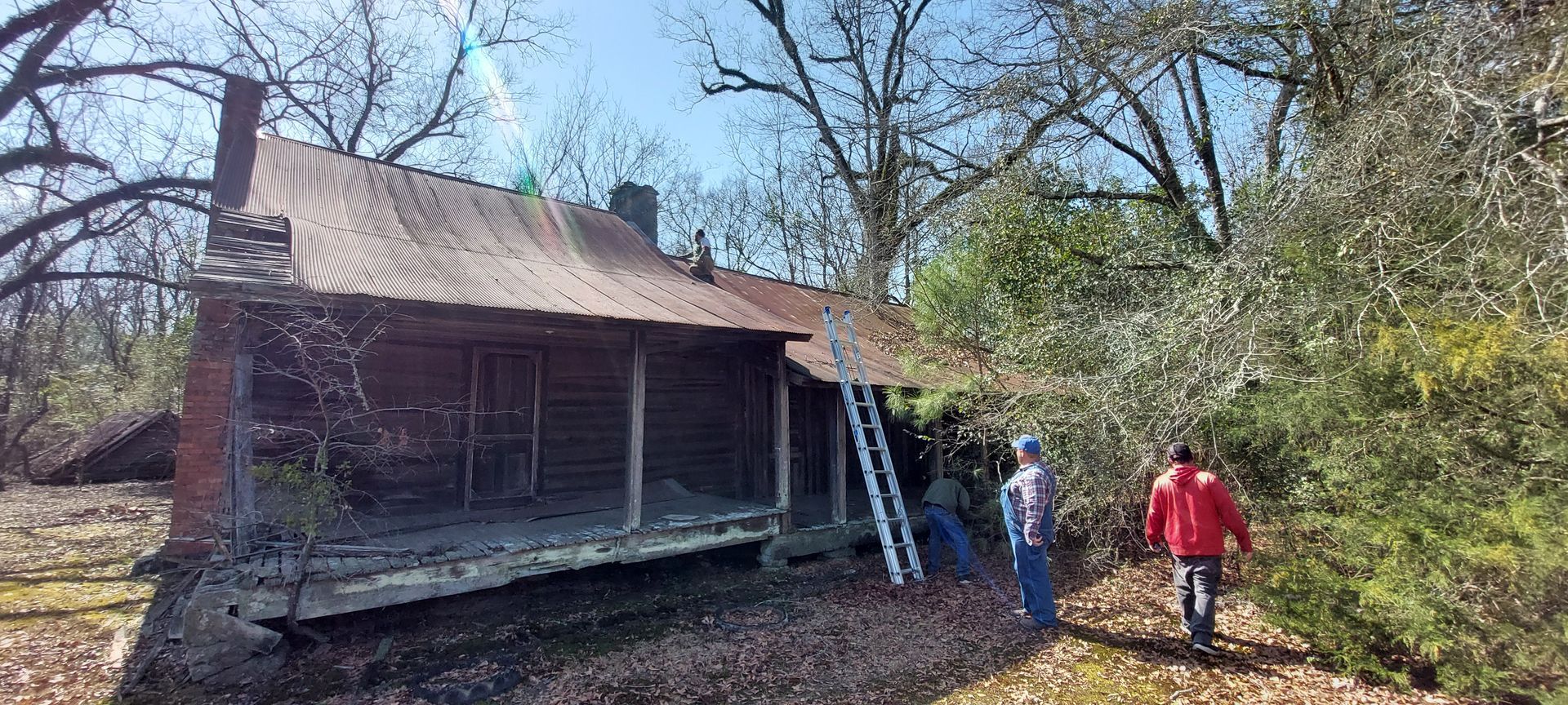Two people walking beside an old weathered cabin in a wooded area on a sunny day