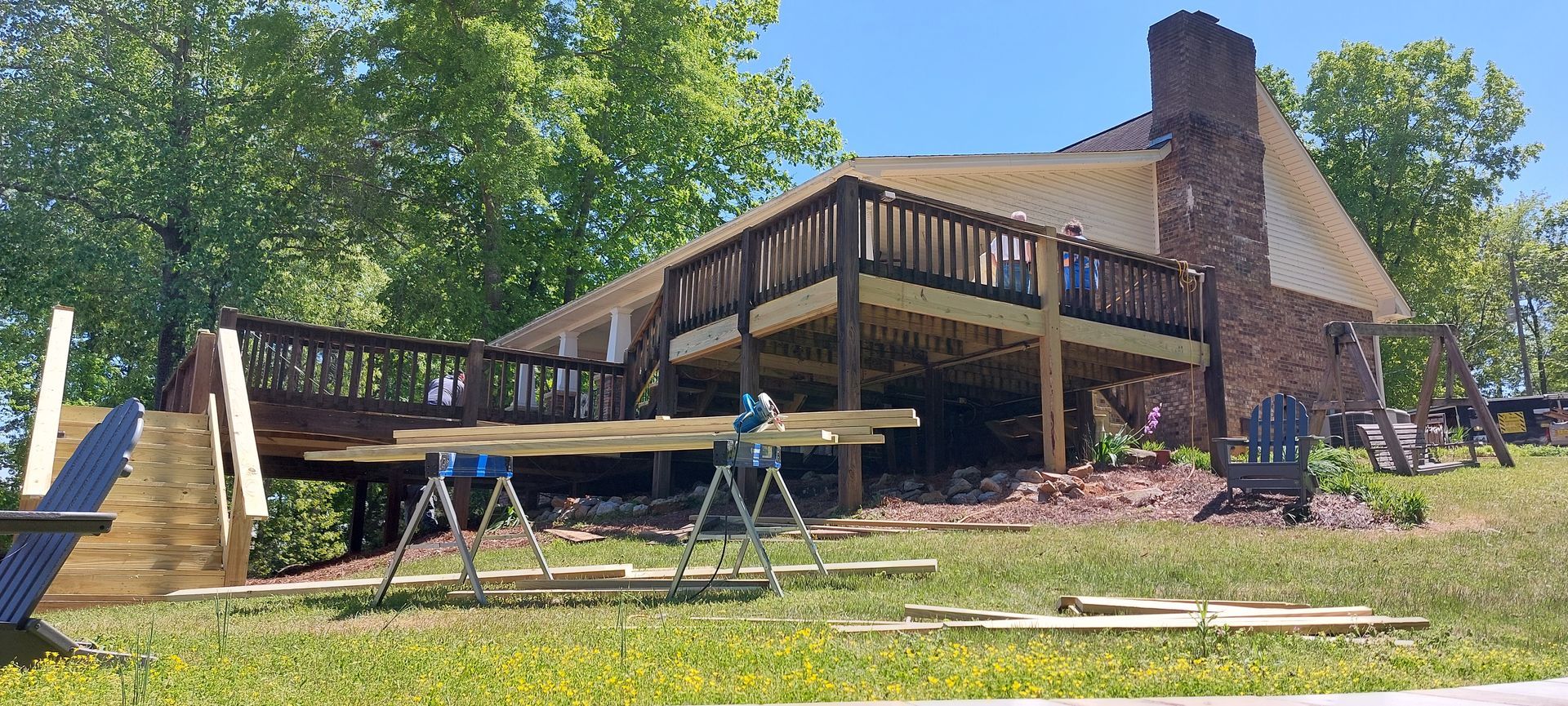 Large wooden deck attached to a brick house, with outdoor tables and chairs in a sunny yard.
