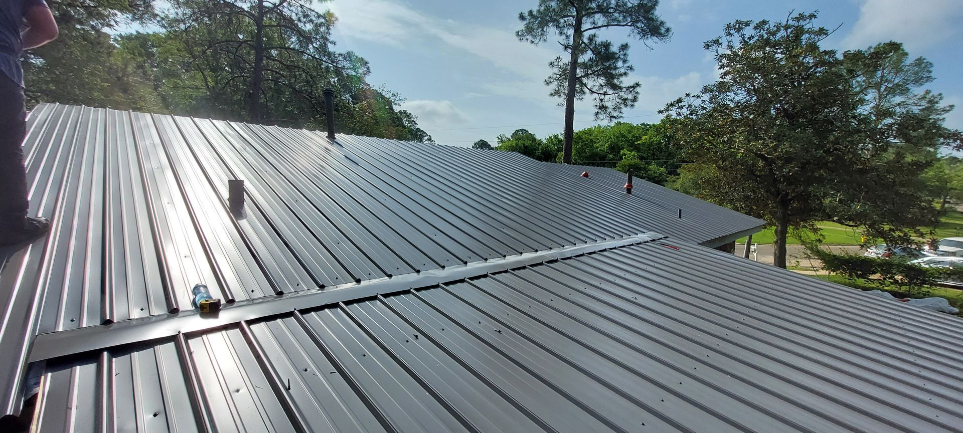 Shiny corrugated metal roof with seams and roof brackets under a blue sky, trees in the background