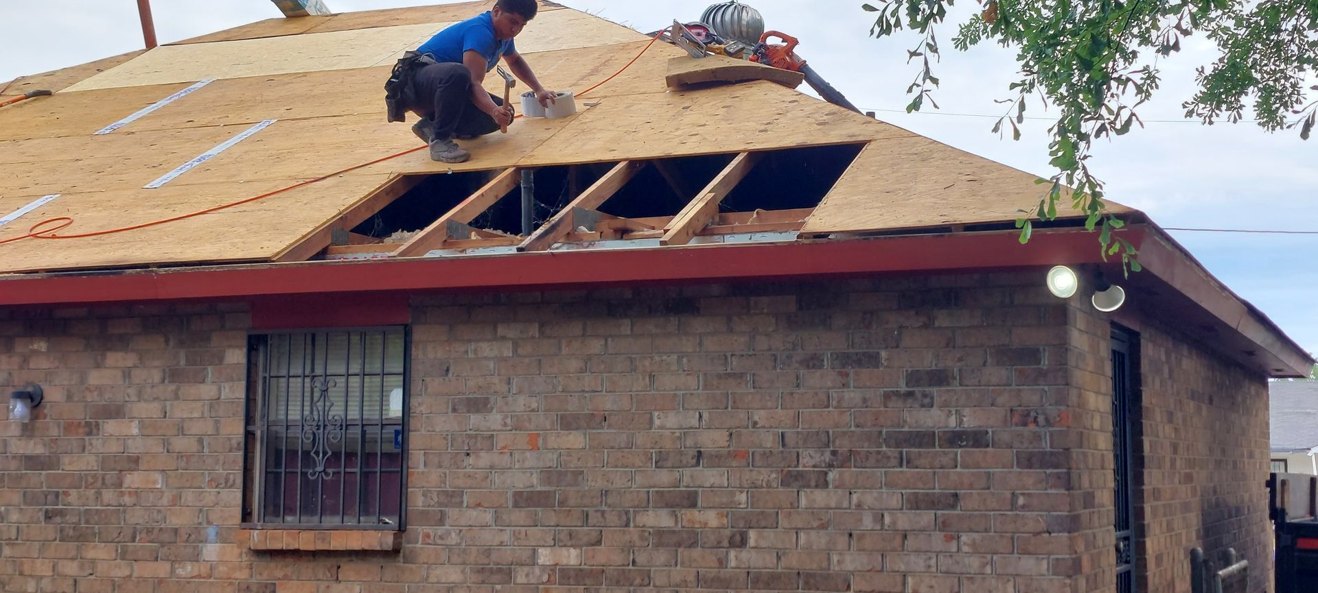 Worker repairing roof on brick house, removing plywood and exposing rafters.