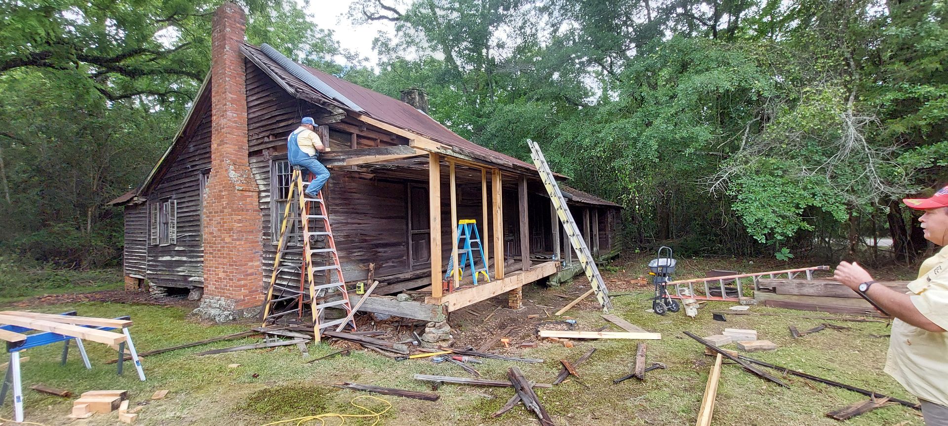 Workers repairing a rustic cabin roof and porch amid fallen branches in a wooded yard