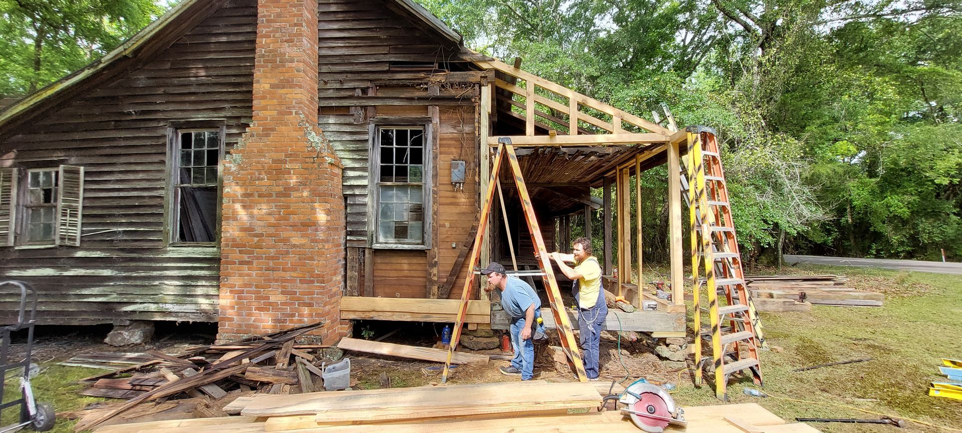 Workers repairing a weathered wooden house with a brick chimney and porch frame removed