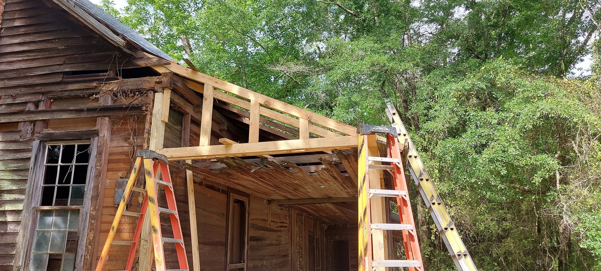 Two ladders beside a rustic wooden house under repair, with exposed roof framing and trees nearby