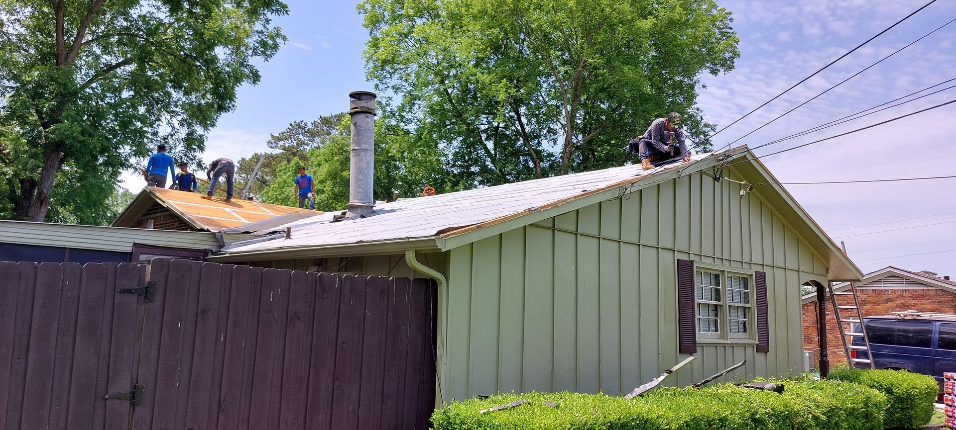 Workers on a green-roofed house repairing roof damage beside a fenced yard
