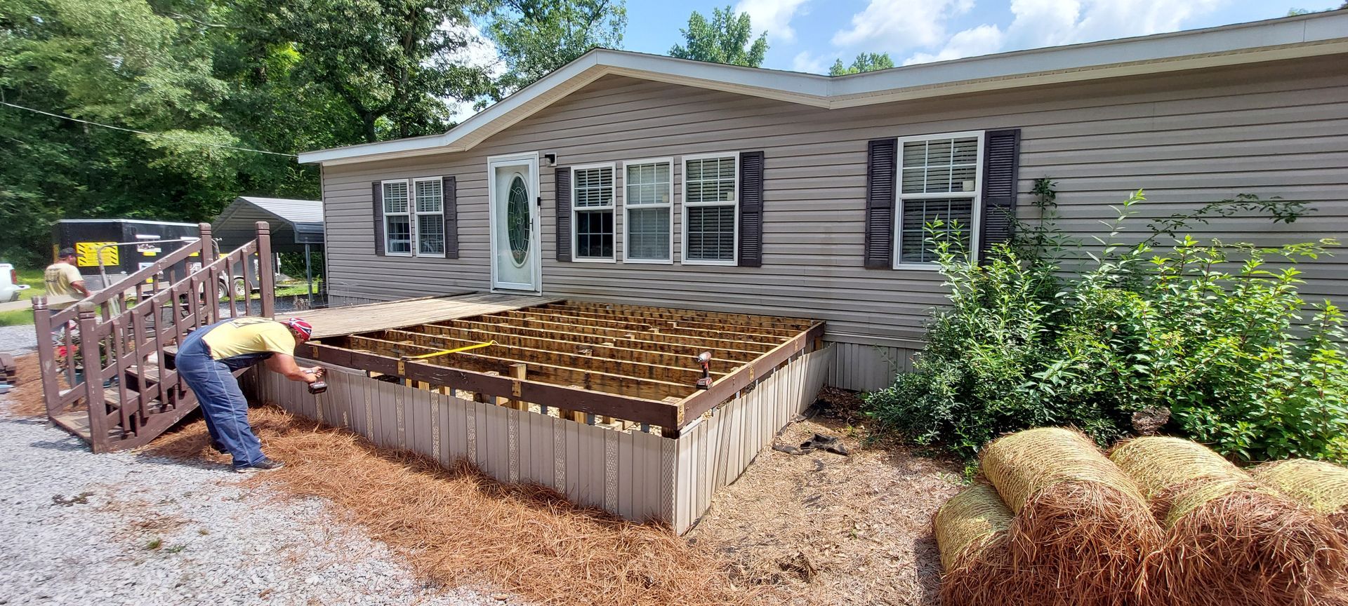 Person installing a raised garden bed beside a gray house, with mulch and stacked materials nearby