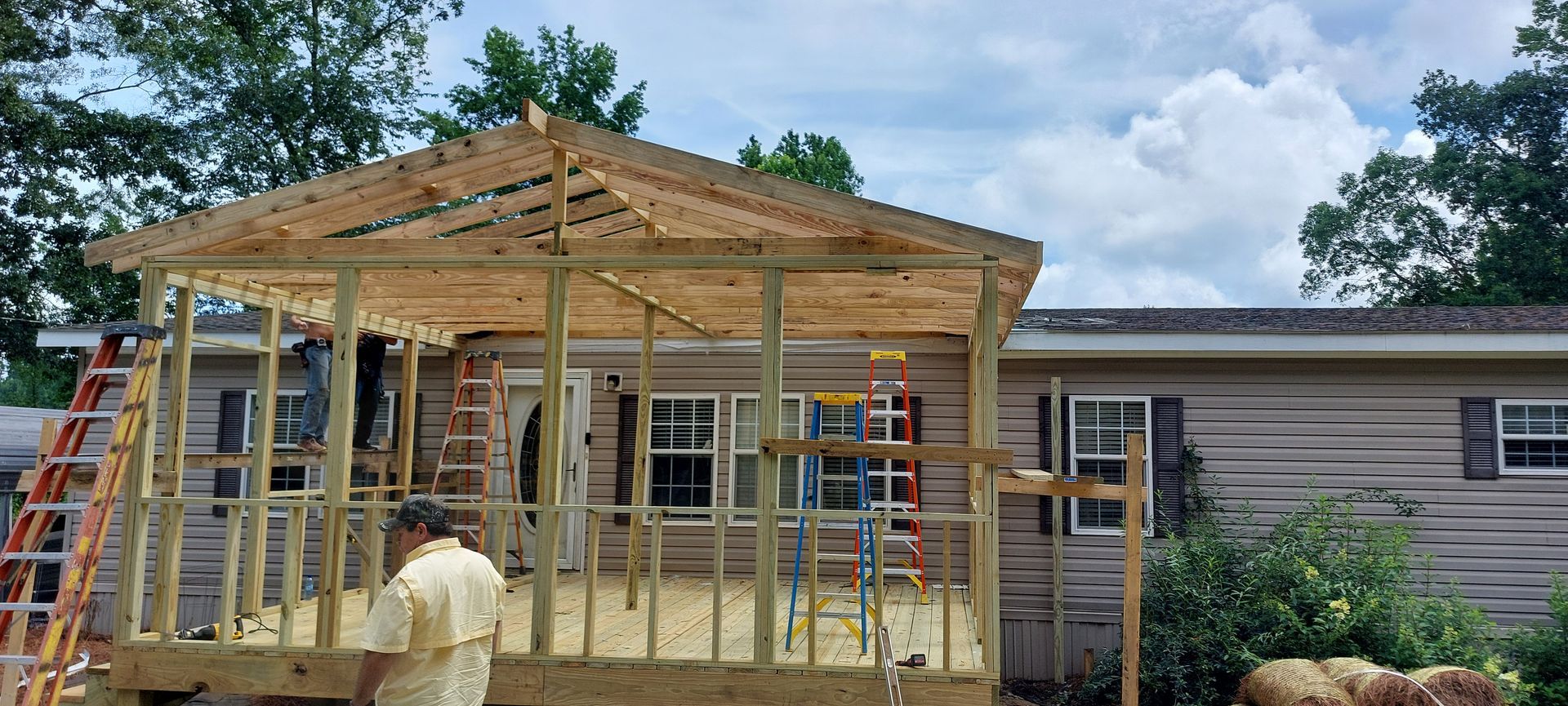 Workers framing a wooden porch roof beside a house, with ladders and unfinished beams.