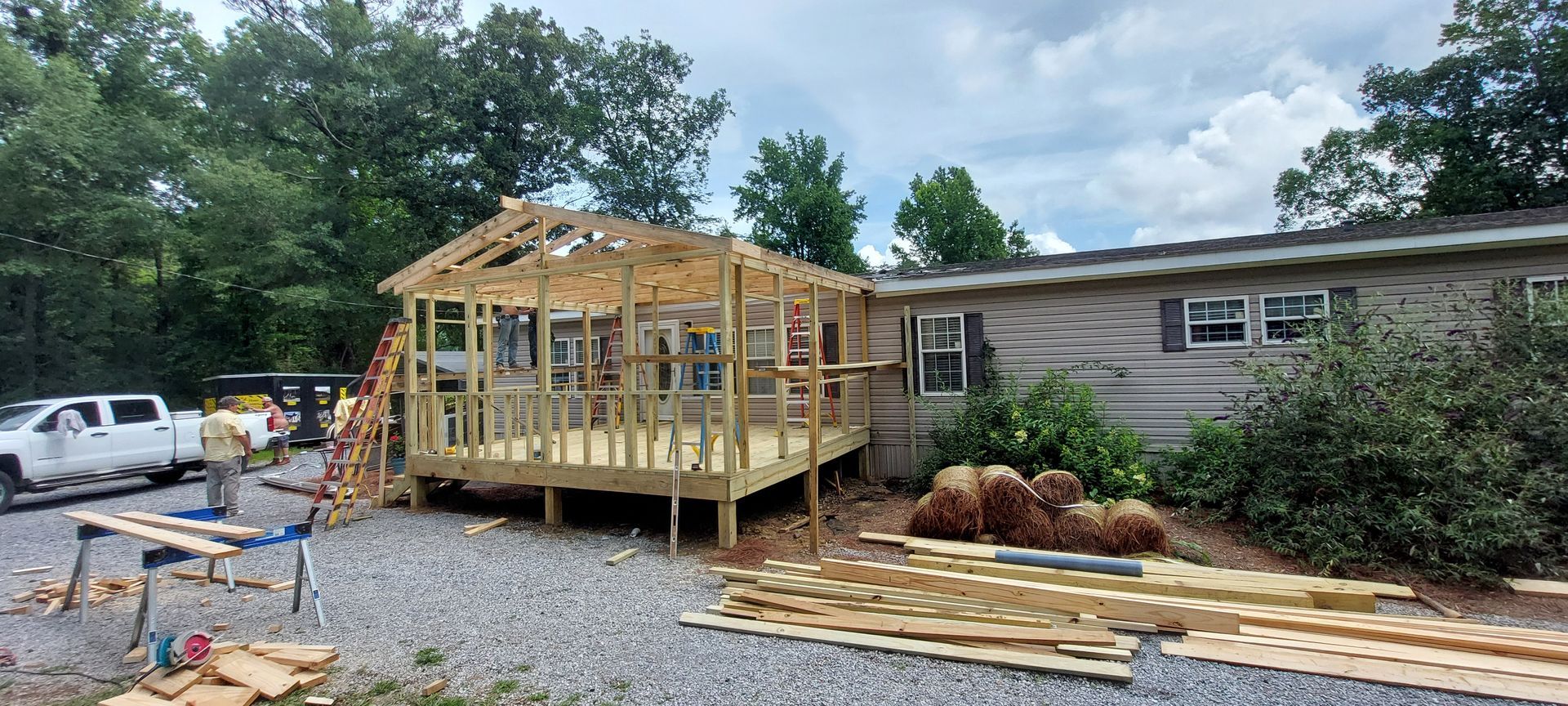 Partially built wooden deck under construction beside a house, with lumber and tools on gravel driveway
