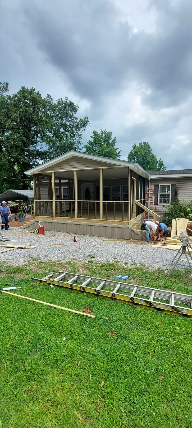 Backyard screened porch under construction with workers, gravel, and tools on a green lawn