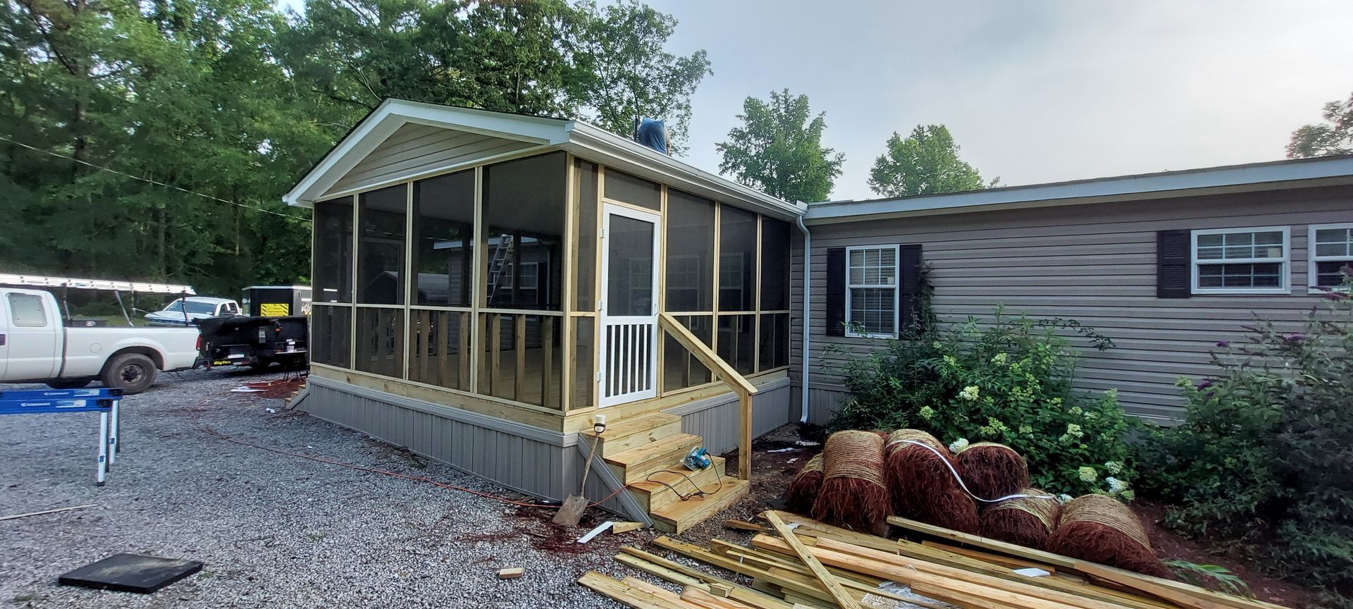 Small screened porch with yellow trim attached to a mobile home, with gravel yard and landscaping.
