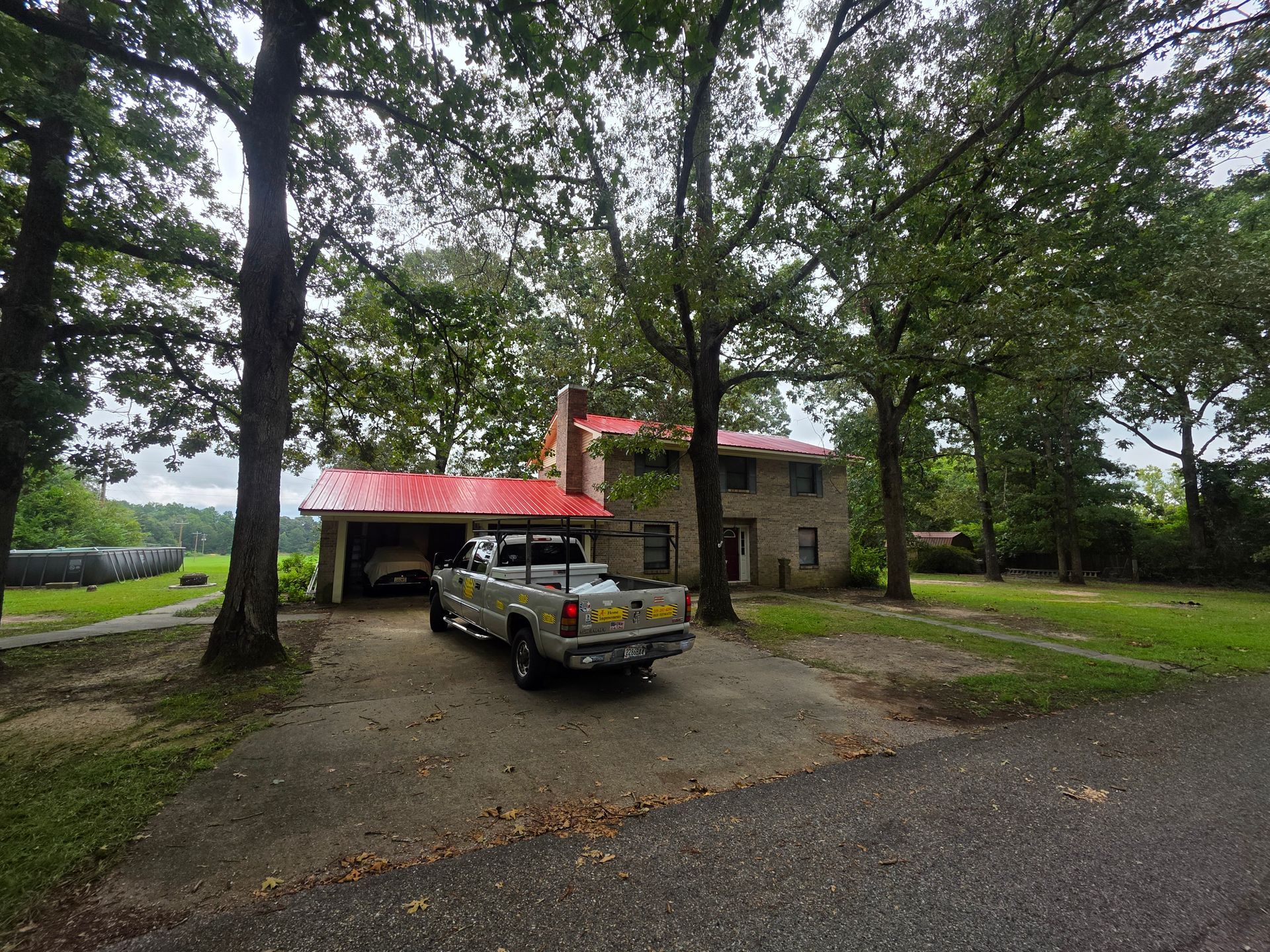 Rural stone house with red roof and pickup truck under large trees on a gravel driveway