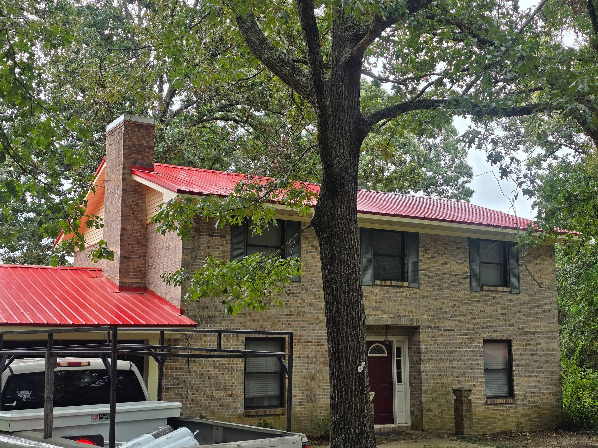 Two-story stone house with red roof and large tree in front, partially covered carport at left