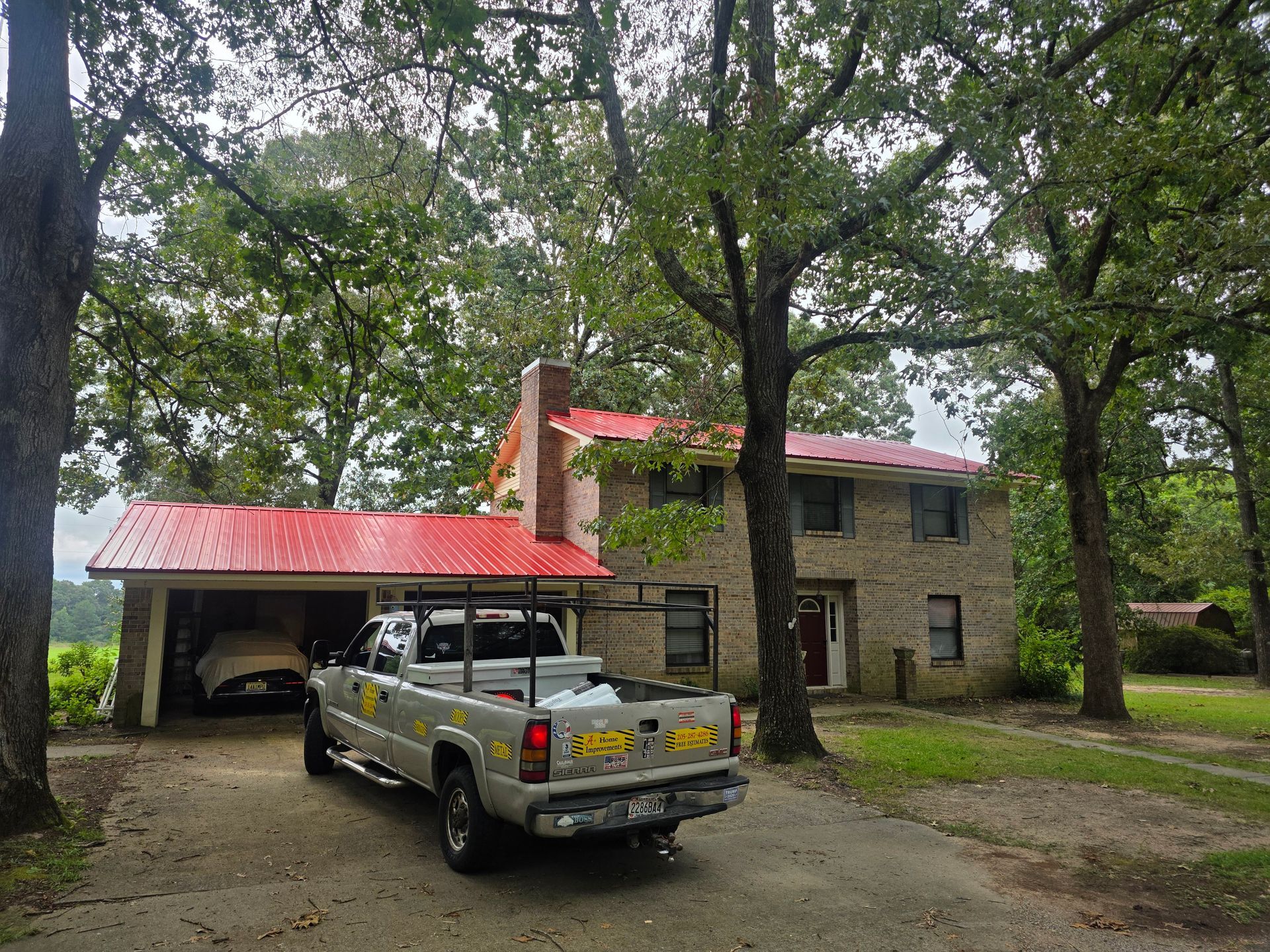 Silver pickup truck parked by a house with a red roof and carport under trees