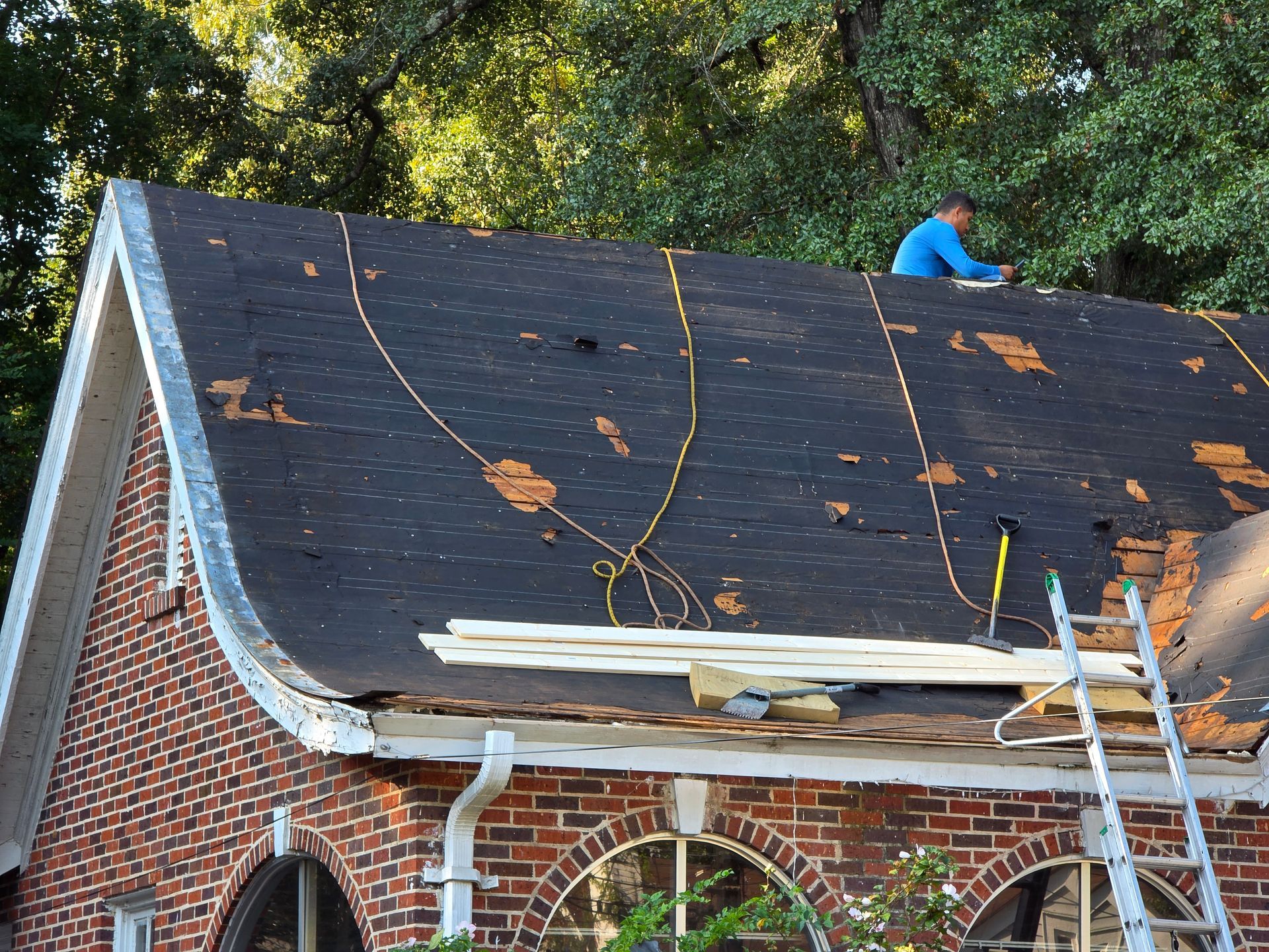 Roof repair on a brick house with black shingles, ladder, and worker on the roof
