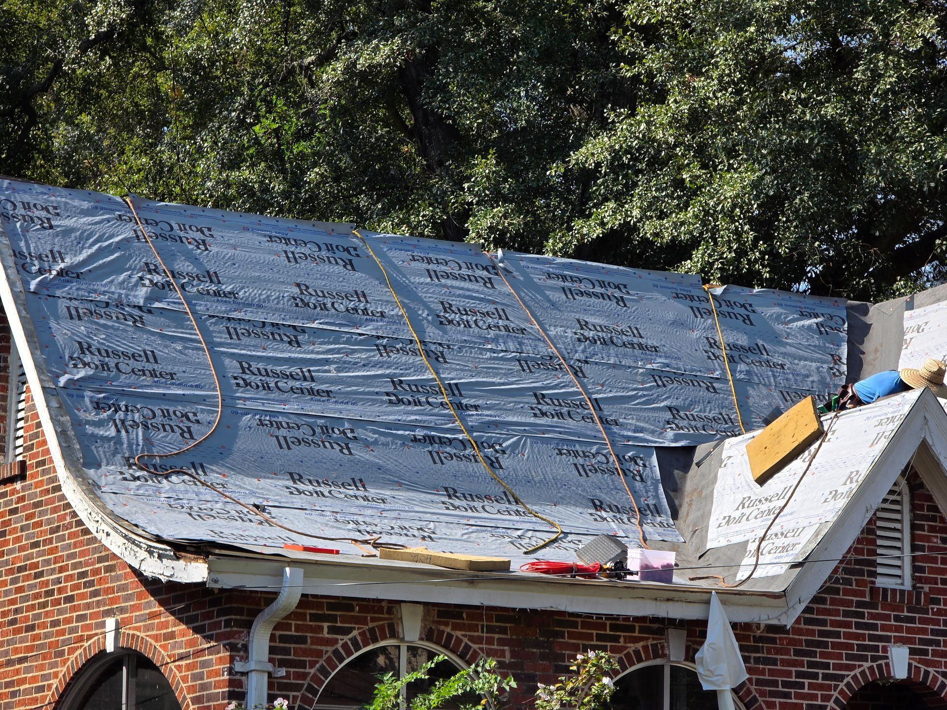 Damaged brick house roof covered with blue tarp and scattered shingles beneath trees