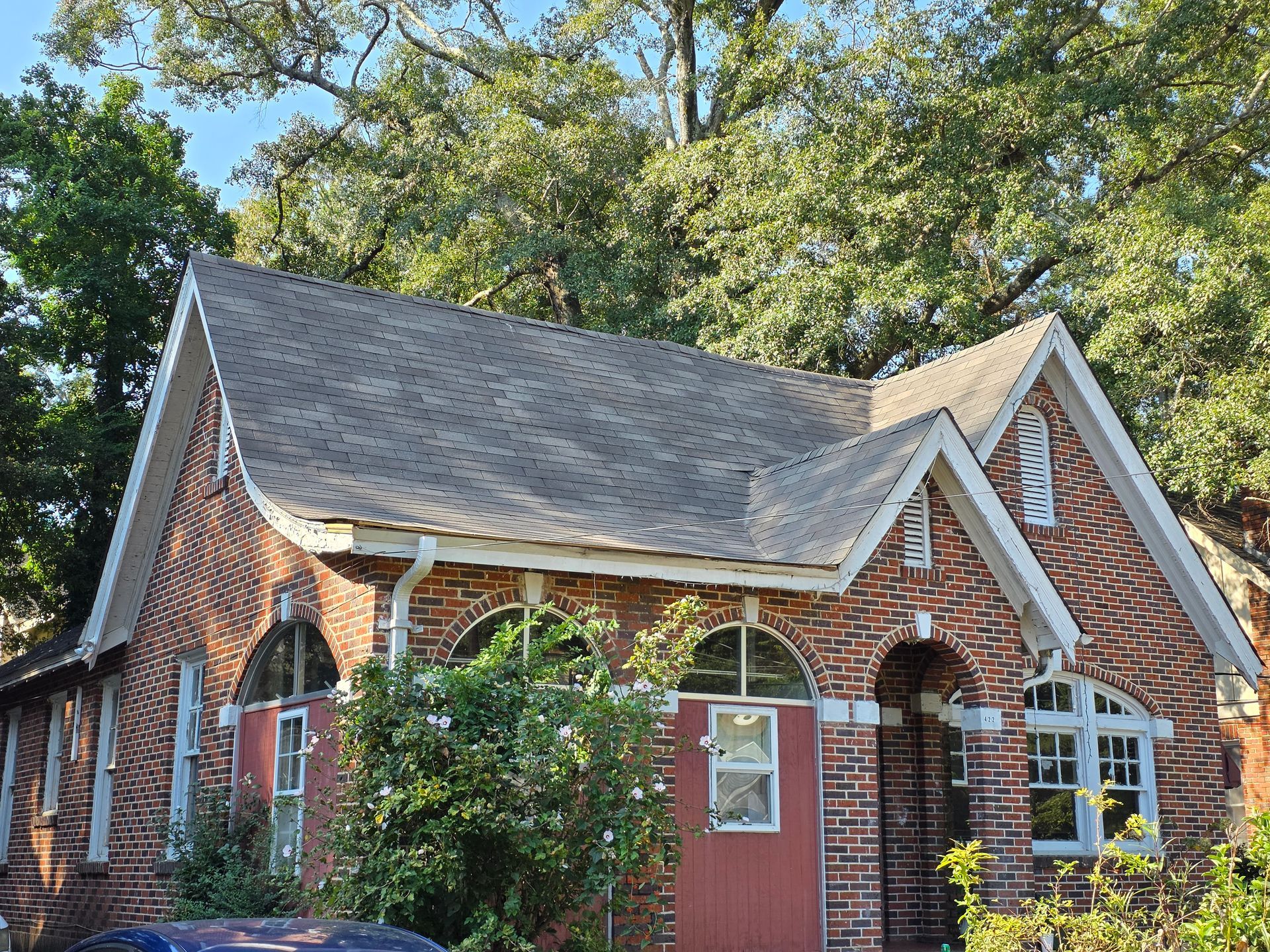 Small red-brick house with a steep gray roof and white-trimmed windows, surrounded by trees