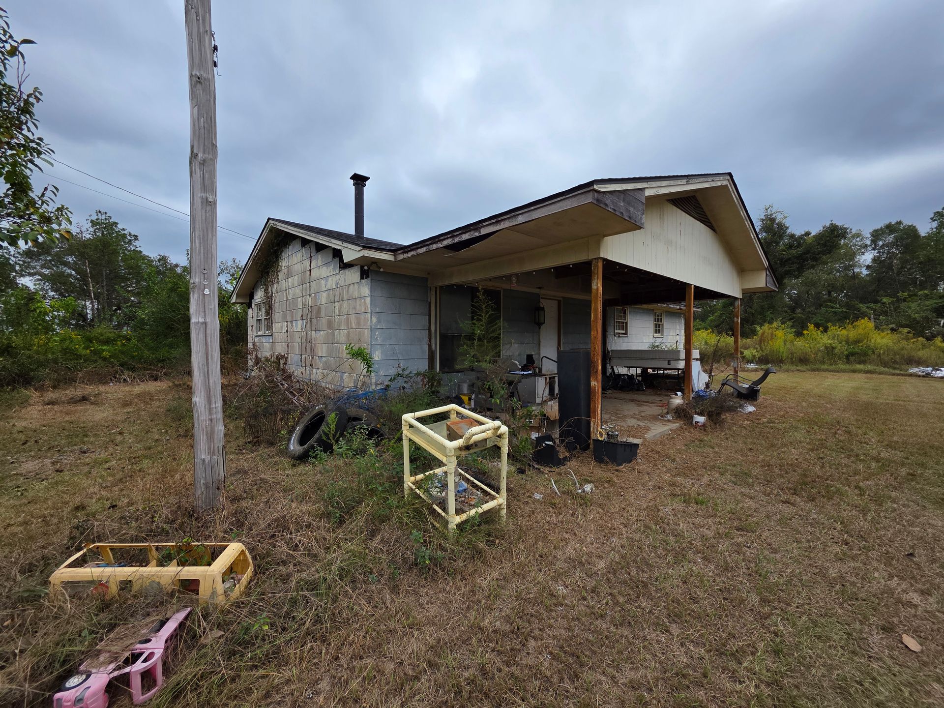Small rural house with a porch, scattered items, and a grassy yard under cloudy skies