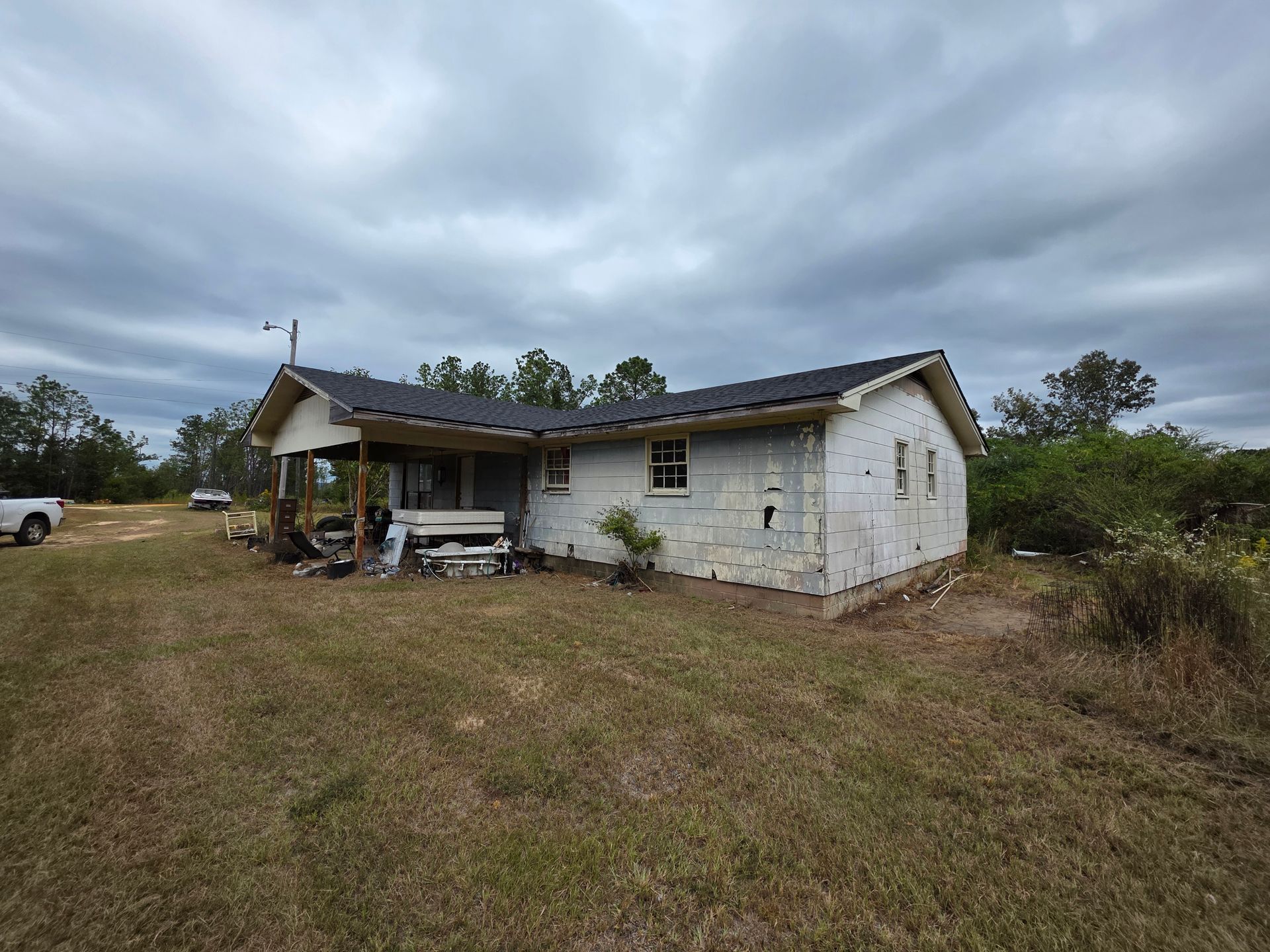 Dilapidated white house on a grassy lot under a cloudy sky, with a small porch and sparse trees nearby