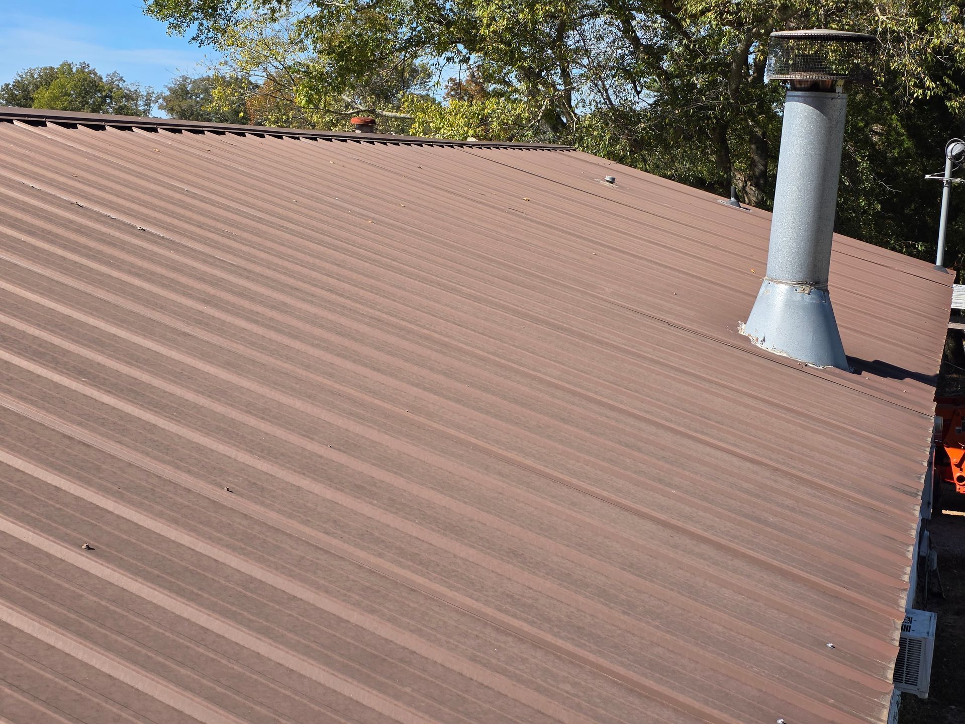 Brown corrugated metal roof with a silver chimney vent under trees