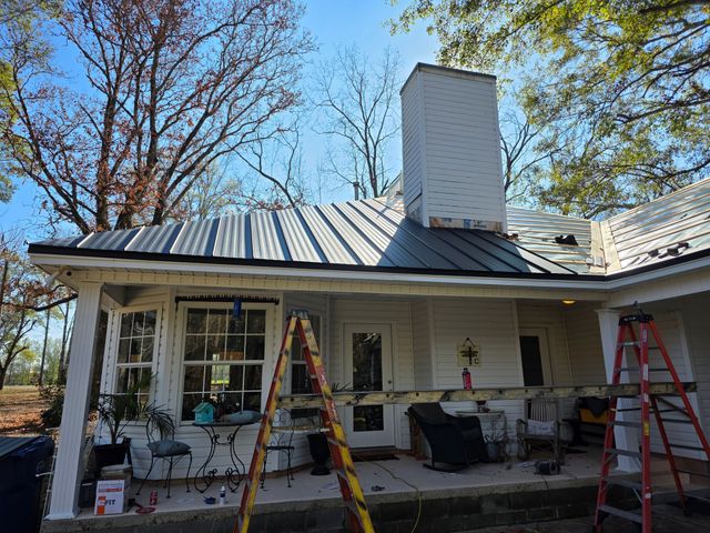 House exterior with a metal roof and chimney, ladders and tools set up for repairs under a covered porch
