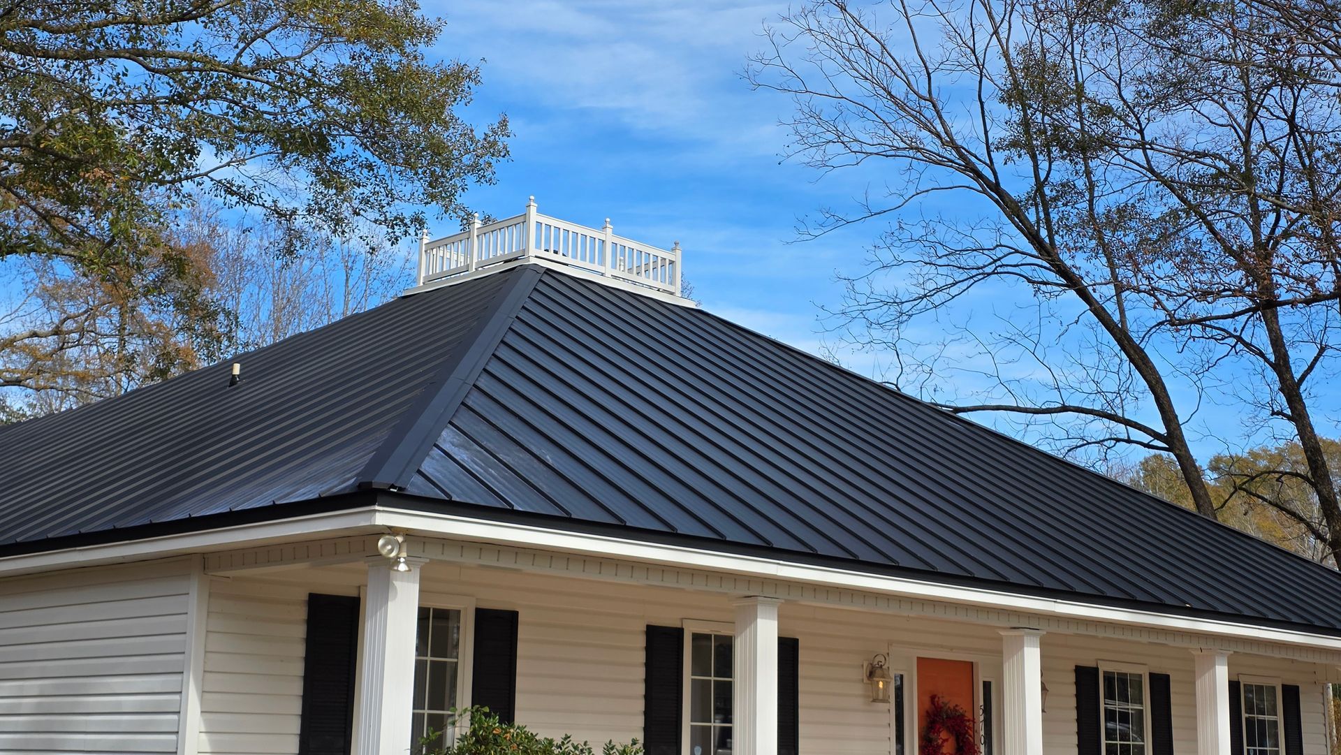 White house with a dark metal roof and dormer against a blue sky, framed by bare trees