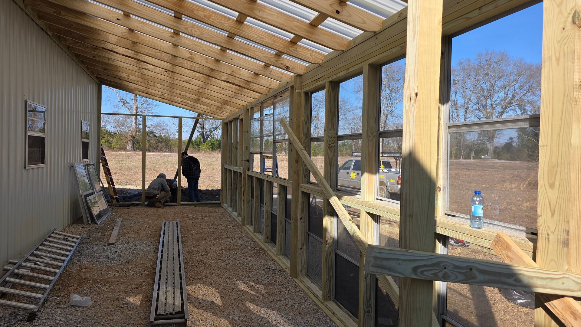 Open porch under construction with lumber piles, wooden framing, and a worker near the back.