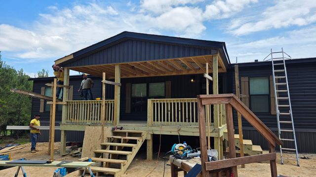 Workers building a wooden front porch and steps on a dark house, with ladders and tools around.