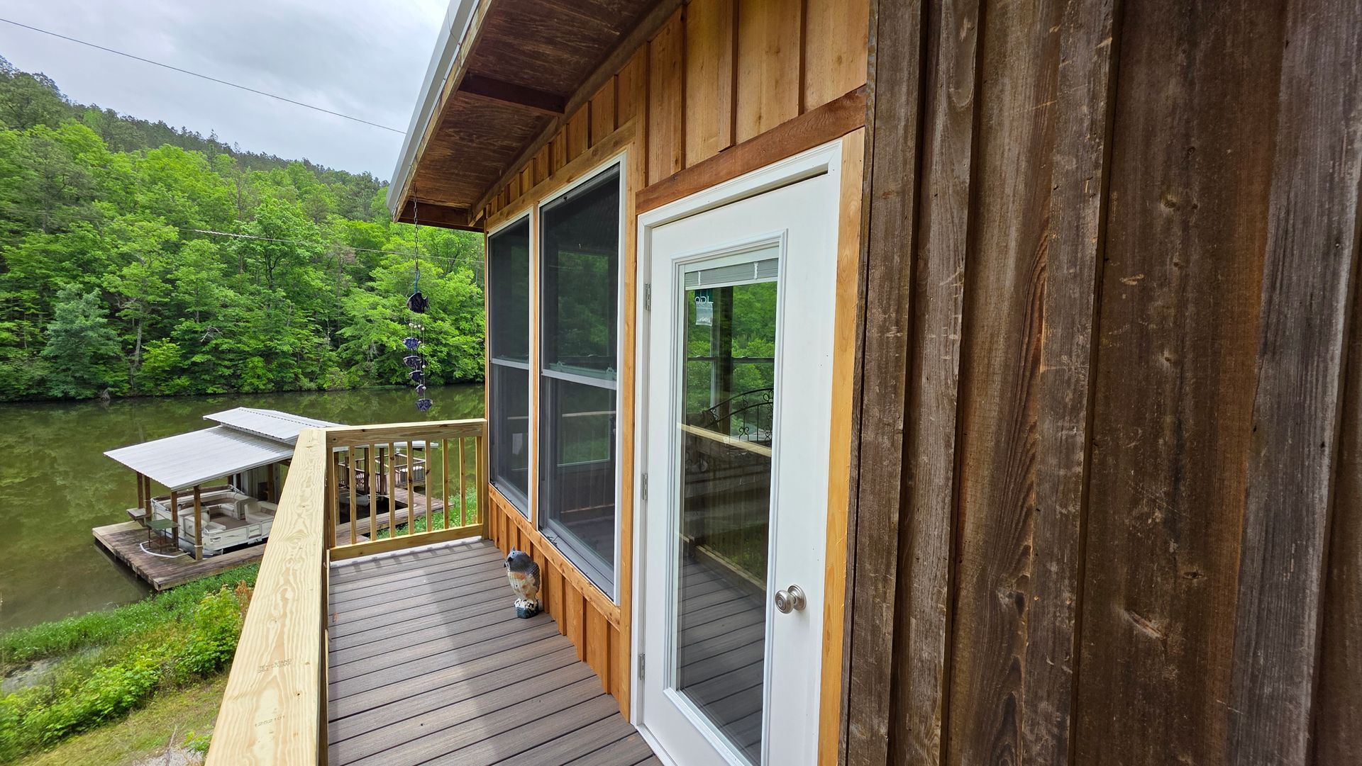 Wooden cabin deck beside a calm lake, with a glass door and green forest beyond.