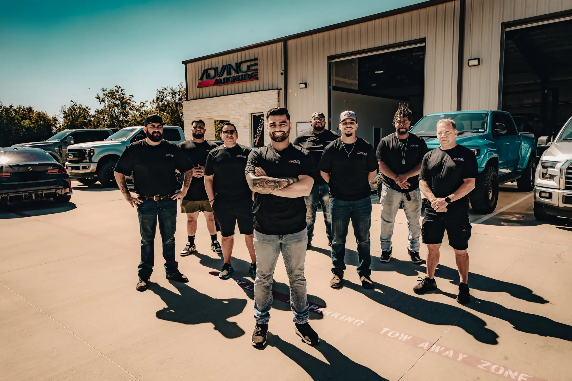 Team of mechanics poses in front of a garage, cars in background. All wearing black shirts, arms crossed. | Advance Automotive