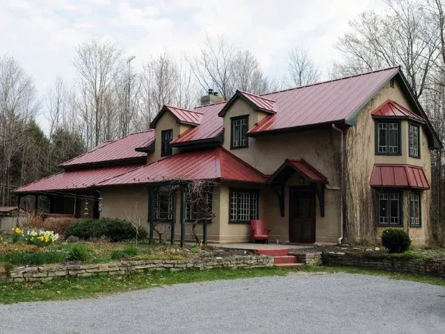 Maison en pierre avec toit en tôle rouge et lucarnes, située au milieu d'arbres dénudés dans une cour boisée.