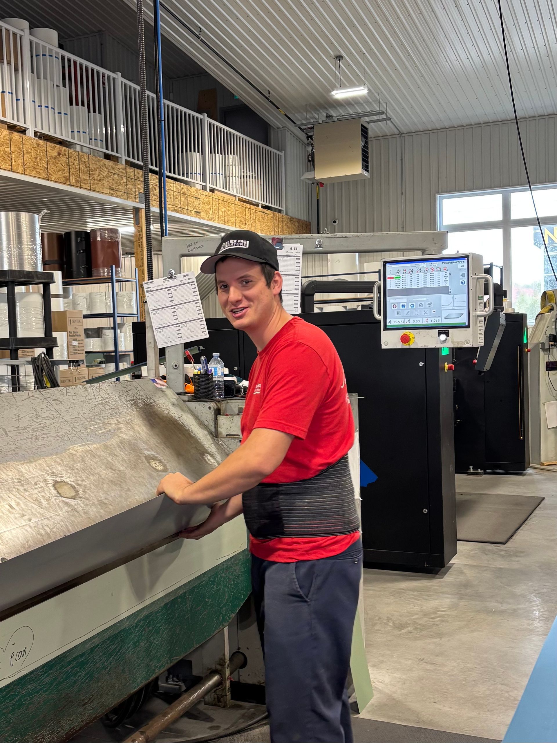 Un ouvrier en chemise et casquette rouges se tient près d'une grande machine industrielle dans un entrepôt.