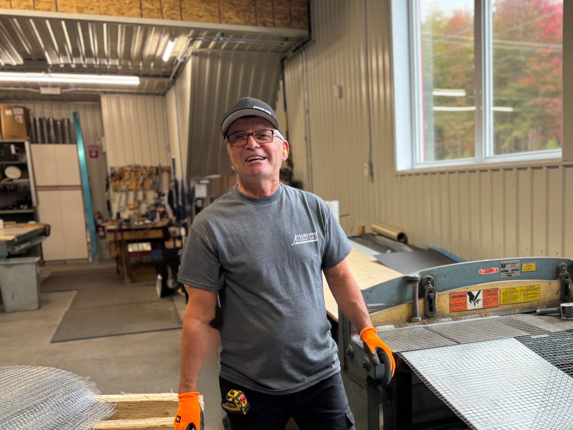 Un homme en chemise grise et gants orange se tient dans un atelier à côté de piles de tôles.