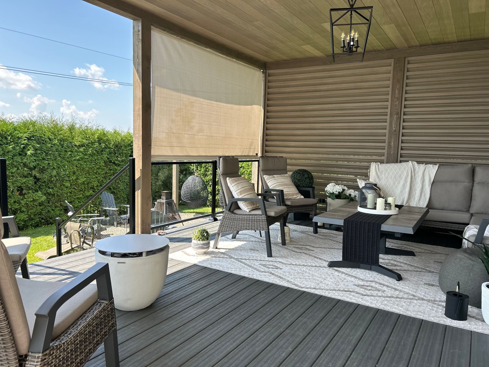 Terrasse couverte avec salon de jardin gris, table ronde, lanterne et verdure extérieure.