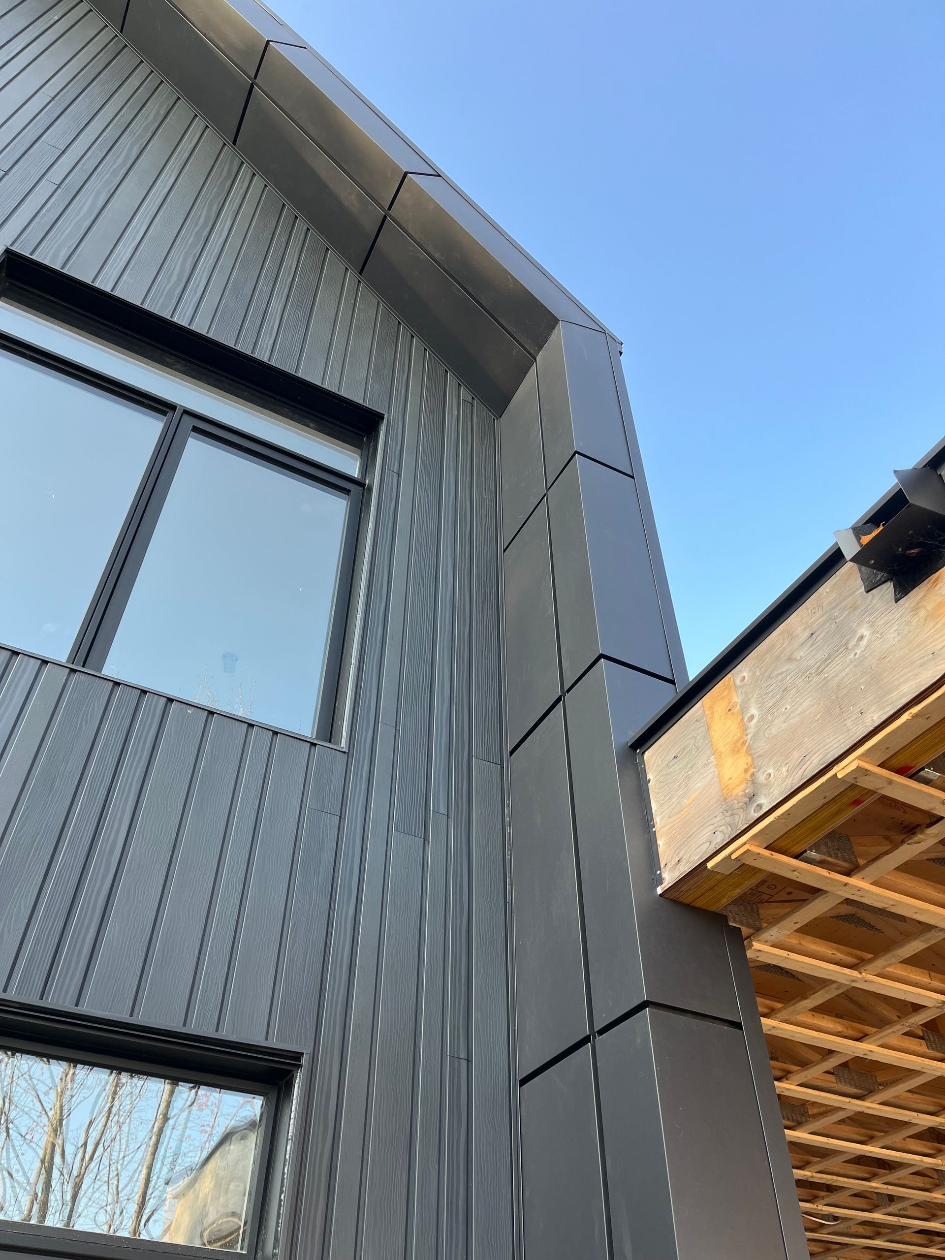 Façade d'un bâtiment moderne gris avec panneaux verticaux et ciel bleu, à côté d'un auvent en bois.